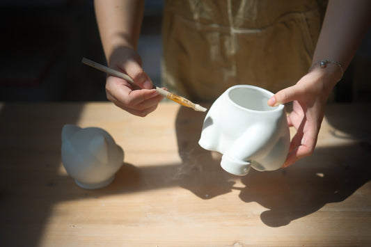 Hands painting a white cat-shaped ceramic urn with a fine brush on a work table